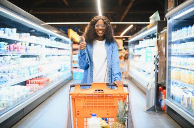 Market alışverişi yapan, alışveriş yapan, yiyecek seçen ve alışveriş yapan mutlu siyah kadın. Modern süpermarketin raflarında market arabasıyla ayakta duruyor..
