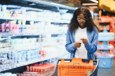 Market alışverişi yapan, alışveriş yapan, yiyecek seçen ve alışveriş yapan mutlu siyah kadın. Modern süpermarketin raflarında market arabasıyla ayakta duruyor..