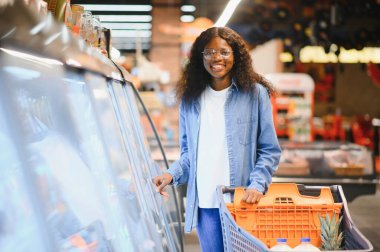 Market alışverişi yapan, alışveriş yapan, yiyecek seçen ve alışveriş yapan mutlu siyah kadın. Modern süpermarketin raflarında market arabasıyla ayakta duruyor..