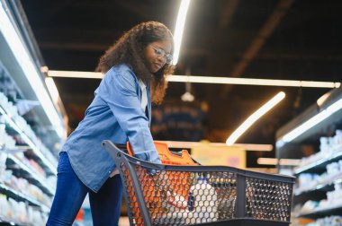 Market alışverişi yapan, alışveriş yapan, yiyecek seçen ve alışveriş yapan mutlu siyah kadın. Modern süpermarketin raflarında market arabasıyla ayakta duruyor..