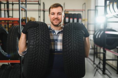 Gülümseyen depo çalışanının portresi yeni araba lastikleri yığınının yanında duruyor..