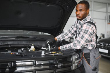 Mühendis otomotiv performansını ve güvenliğini sağlamak için gelişmiş mekanik araçlar kullanarak otomobil motorunu inceliyor. Afrikalı Amerikalı garaj çalışanı yıllık araç kontrolü yapıyor..