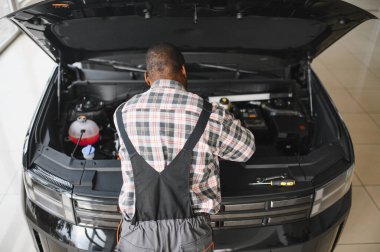 Mühendis otomotiv performansını ve güvenliğini sağlamak için gelişmiş mekanik araçlar kullanarak otomobil motorunu inceliyor. Afrikalı Amerikalı garaj çalışanı yıllık araç kontrolü yapıyor..