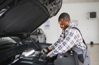 Mühendis otomotiv performansını ve güvenliğini sağlamak için gelişmiş mekanik araçlar kullanarak otomobil motorunu inceliyor. Afrikalı Amerikalı garaj çalışanı yıllık araç kontrolü yapıyor..