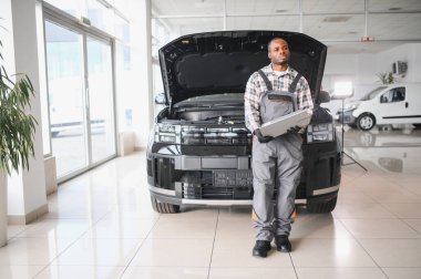 Black auto mechanic running diagnostic on laptop while analyzing engine problems in a workshop.