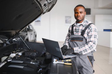 Mühendis otomotiv performansını ve güvenliğini sağlamak için gelişmiş mekanik araçlar kullanarak otomobil motorunu inceliyor. Afrikalı Amerikalı garaj çalışanı yıllık araç kontrolü yapıyor..