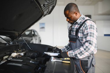 Tamirci, otomobil tamirhanesinde pano ile arabayı dikkatlice inceliyor. Odaklanmış profesyonel araç bakım ve kayıt ayrıntıları.