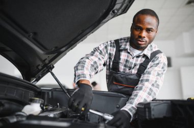 Mühendis otomotiv performansını ve güvenliğini sağlamak için gelişmiş mekanik araçlar kullanarak otomobil motorunu inceliyor. Afrikalı Amerikalı garaj çalışanı yıllık araç kontrolü yapıyor..