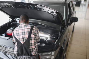 Mühendis otomotiv performansını ve güvenliğini sağlamak için gelişmiş mekanik araçlar kullanarak otomobil motorunu inceliyor. Afrikalı Amerikalı garaj çalışanı yıllık araç kontrolü yapıyor..