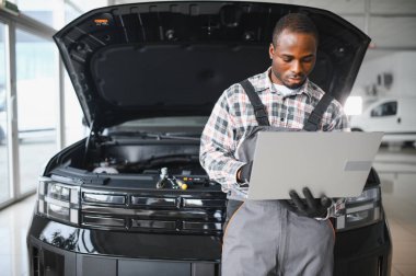 Black auto mechanic running diagnostic on laptop while analyzing engine problems in a workshop.