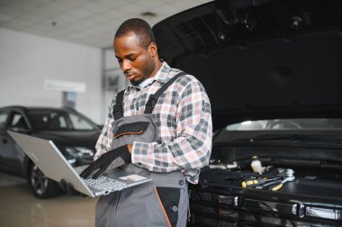 Black auto mechanic running diagnostic on laptop while analyzing engine problems in a workshop.
