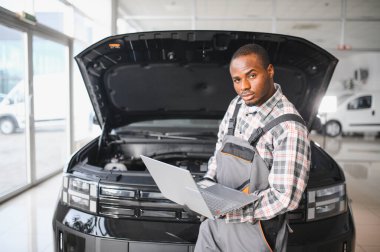 Mühendis otomotiv performansını ve güvenliğini sağlamak için gelişmiş mekanik araçlar kullanarak otomobil motorunu inceliyor. Afrikalı Amerikalı garaj çalışanı yıllık araç kontrolü yapıyor..