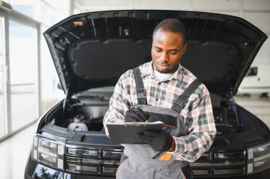 Tamirci, otomobil tamirhanesinde pano ile arabayı dikkatlice inceliyor. Odaklanmış profesyonel araç bakım ve kayıt ayrıntıları.