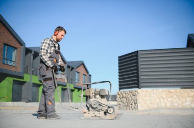 Male Worker using vibratory plate compactor to firm soil at worksite. builder. After laying of paving slabs, compacts paving stones with a gasoline vibratory compactor for subsequent. Safety at work clipart