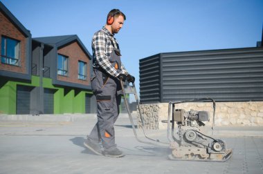 Erkek işçi, inşaat alanında toprak sıkıştırmak için titreşimli plaka sıkıştırıcısı kullanıyor. İnşaatçı. Döşeme levhaları döşedikten sonra, kompakt taşları akabinde kullanılacak olan benzinli titreşim sıkıştırıcı ile kaplıyor..