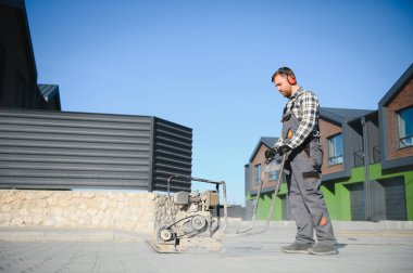 Erkek işçi, inşaat alanında toprak sıkıştırmak için titreşimli plaka sıkıştırıcısı kullanıyor. İnşaatçı. Döşeme levhaları döşedikten sonra, kompakt taşları akabinde kullanılacak olan benzinli titreşim sıkıştırıcı ile kaplıyor..