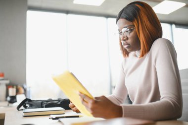 Genç Afrikalı Amerikalı öğrenci üniversitede okuyor, araştırma gazetesinde çalışıyor, açık kitaplarla masada oturuyor..