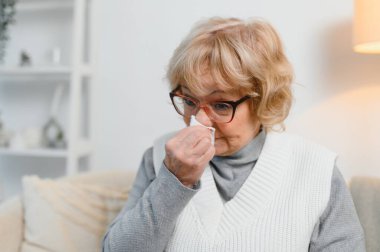 Yaşlı hasta kadın oturma odasında kanepede oturmuş sıcak ekoseyle kaplanmış hapşırıyor kağıt peçete tutuyor burnu akıyor sağlıksız, mevsimsel soğuk, zayıf bağışıklık sistemi konsepti.