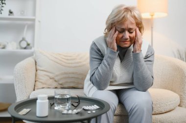 Evdeki kanepede oturan yaşlı kadın, baş ağrısı, ağrı ve kendini kötü hissetme ifadesi ile.