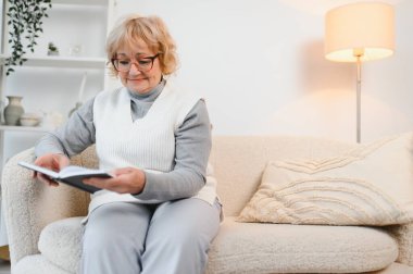 Kanepede oturmuş evde kitap okuyan neşeli, son sınıf öğrencisi bir kadın. Eğitim, olgun, boş zaman kavramı.