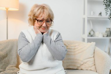 Olgun bir kadın kanepede oturuyor, iltihaplı lenf bezlerine dokunuyor, rahatsızlık ve boğaz ağrısı hissediyor. Solunum yolu enfeksiyonu ya da kronik bademcik iltihabı olan hasta bir kadın..