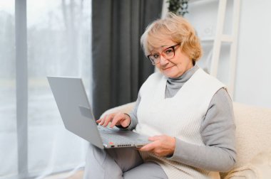 Üst düzey kadın dizüstü bilgisayar kullanıyor, evdeki kanepede oturuyor..