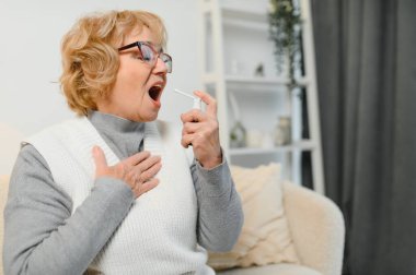 Bir kadın, bir dizi mendille çevriliyken rahatlatıcı bir boğaz spreyi kullanarak kanepesine rahatça yerleşir. Boğaz rahatsızlığını hafifletmek için ideal bir ev yapımı ilaç..