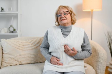 Yatağında oturan yaşlı kadının midesi ağrıyor.,