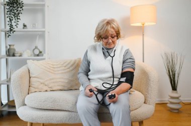 Yüksek tansiyon. Olgun kadın kan basıncını evde tonometreyle ölçüyor, boş alanı olan bir panorama..