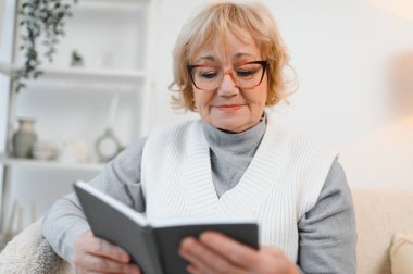 Kanepede oturmuş evde kitap okuyan neşeli, son sınıf öğrencisi bir kadın. Eğitim, olgun, boş zaman kavramı.