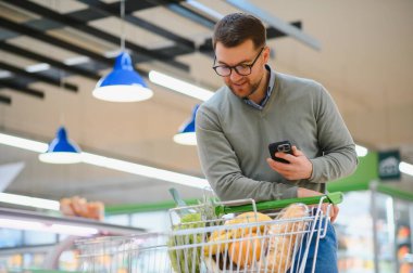 Süpermarketten alışveriş yaparken cep telefonu uygulaması kullanan mutlu adam..