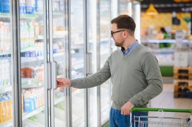Süpermarkette alışveriş yaparken akıllı telefon kullanan ve kameraya bakan mutlu bir adam..