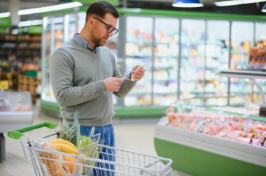 Şok olmuş zarif bir adamın portresi süpermarketten yiyecek alırken elinde uzun bir alışveriş listesi tutuyor..