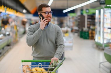 Süpermarketten alışveriş yaparken cep telefonu uygulaması kullanan mutlu adam..