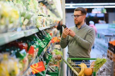 Süpermarketten alışveriş yaparken cep telefonu uygulaması kullanan mutlu adam..