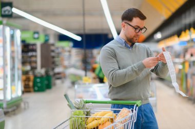 Şok olmuş, yuvarlak gözlü beyaz bir adam son derece uzun bir banknota bakıyor. Market alışverişi yapan ve fiyatlara şaşıran harika bir alıcı..