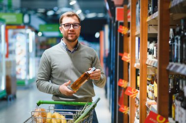 Süpermarketin alkol reyonunda duran sert içkileri seçen ve raftan viski alan bir adamın portresi. Alıcı süpermarketten alkol alır.