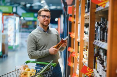 Bir adam süpermarketin rafından alkollü içecekler alır. Alkol almak için markete gidiyorum..