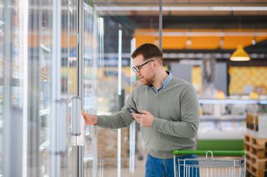 Süpermarketten alışveriş yaparken cep telefonu uygulaması kullanan mutlu adam..
