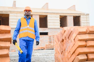 Afrika kökenli Amerikalı bir inşaat işçisi, iş kıyafetleri ve sıkı bir şapkayla tuğla bir ev inşa ediyor..