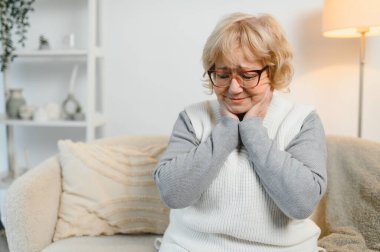 Olgun bir kadın kanepede oturuyor, iltihaplı lenf bezlerine dokunuyor, rahatsızlık ve boğaz ağrısı hissediyor. Solunum yolu enfeksiyonu ya da kronik bademcik iltihabı olan hasta bir kadın..