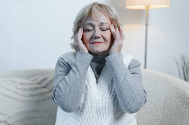 Evdeki kanepede oturan yaşlı kadın, baş ağrısı, ağrı ve kendini kötü hissetme ifadesi ile.