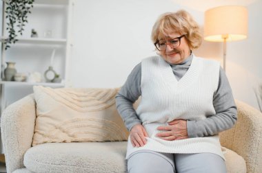 Yatağında oturan yaşlı kadının midesi ağrıyor.,