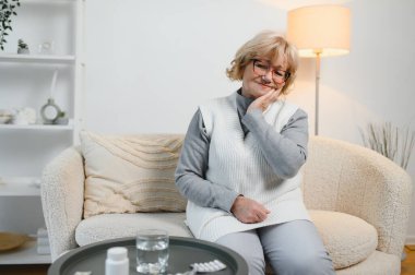 Kanepede oturan yaşlı bir kadın ciddi diş ağrısı, yaşlılıkta sağlık sorunları, yaşam kalitesi düşük. Gri saçlı büyükanne çenesini tutuyor.