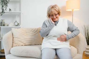 Yatağında oturan yaşlı kadının midesi ağrıyor.,