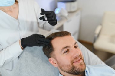 Yakışıklı bir adamın kuaförde saç bakımına gidişinin yan görüntüsü. Adam estetik kliniğinde mezoterapi görüyor, terapist eldiven takıyor kafa derisine iğne yapıyor, yakın çekim yapıyor.