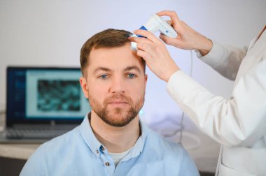 Kadın doktor dermatoskopla bir adamın kafa derisine bakıyor. Bir tricholog tarafından kafa derisi hastalığı teşhisi konuldu. Köstebek muayenesi, kellik, saç nakli..