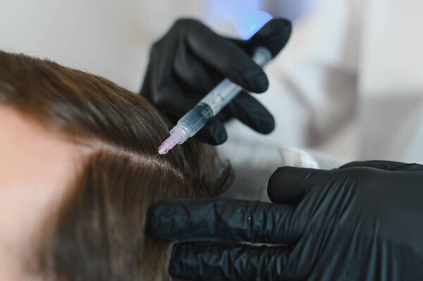 Мужчина получает лечение волос в салоне красоты, проводит сеанс мезотерапии. Женские руки терапевта в защитных перчатках делают инъекцию шприца.