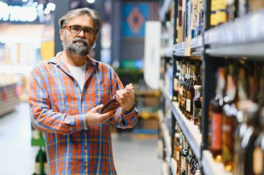 Sakallı bir adam elinde bir şişe konyakla süpermarkette duruyor..