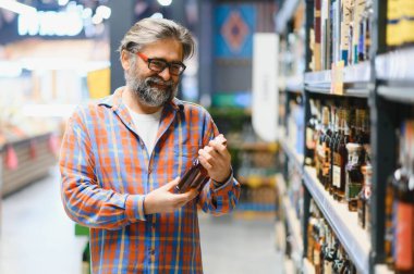 Sakallı bir adam elinde bir şişe konyakla süpermarkette duruyor..